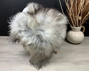 Può includere: Un soffice tappeto in pelle di pecora grigio e bianco. Il tappeto ha una lunga e morbida pelliccia ed è posto su una superficie di legno chiaro. Sullo sfondo, un vaso bianco con un manico marrone contiene rami secchi.
