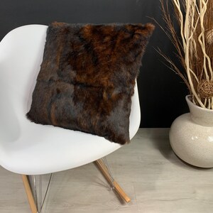 May include: A square decorative pillow with a brown and black fur pattern rests on a white chair. The pillow's texture contrasts with the smooth chair. A vase with decorative branches and orbs is in the background.