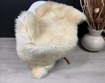 May include: A plush, cream-colored sheepskin rug with long, dense fur fibers. The rug features an irregular, natural shape with a soft, thick texture that covers the seat of a chair, showcasing a uniform off-white color throughout the piece.