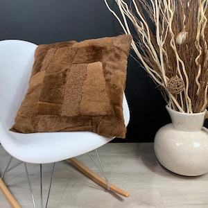 Può includere: Un cuscino decorativo quadrato marrone con un design patchwork poggia su una sedia a dondolo bianca. Il cuscino ha un aspetto morbido e strutturato. Un vaso di ceramica bianco con manico in corda contiene rami decorativi.