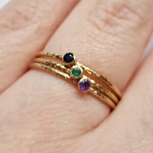 May include: Three gold rings stacked on a finger, each with a small, round gemstone in black, green, and purple. The rings have a textured surface. The finger is in focus against a blurred background.