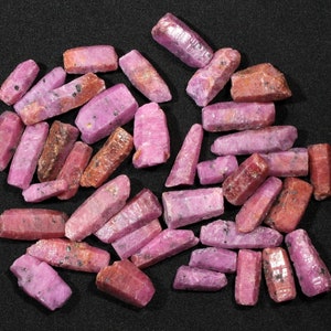 May include: A collection of pink and red ruby crystals, some with a rough texture, arranged on a black surface.