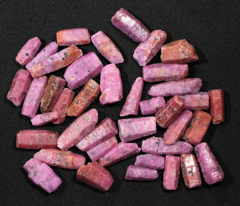May include: A collection of pink and red ruby crystals, some with a rough texture, arranged on a black surface.