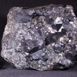 May include: A close-up of a dark gray rock with black crystals. The crystals are octahedral in shape and have a metallic luster. The rock is about 1 cm in size.