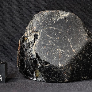 May include: A large, dark, irregularly shaped stone with a rough, textured surface. The stone is black with some lighter areas and a few visible cracks. A black cube with "T. 1 CM" printed on it is next to the stone.