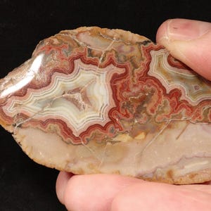 Puede incluir: Una rebanada de piedra de ágata pulida con intrincados patrones de colores rojo, blanco y marrón. La piedra se sostiene en una mano, mostrando su belleza natural y formaciones geológicas únicas. Las capas de la ágata crean un diseño orgánico visualmente atractivo.