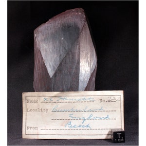 May include: A dark gray, metallic, irregularly shaped rock with a label that reads "X. Hematite No. 232 Locality Cumberland England From Beoch". A 1 cm ruler is included for scale.