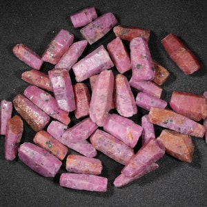 May include: A collection of pink ruby crystals, each with a pointed tip and a rough, natural texture. The crystals are arranged in a pile on a black background.