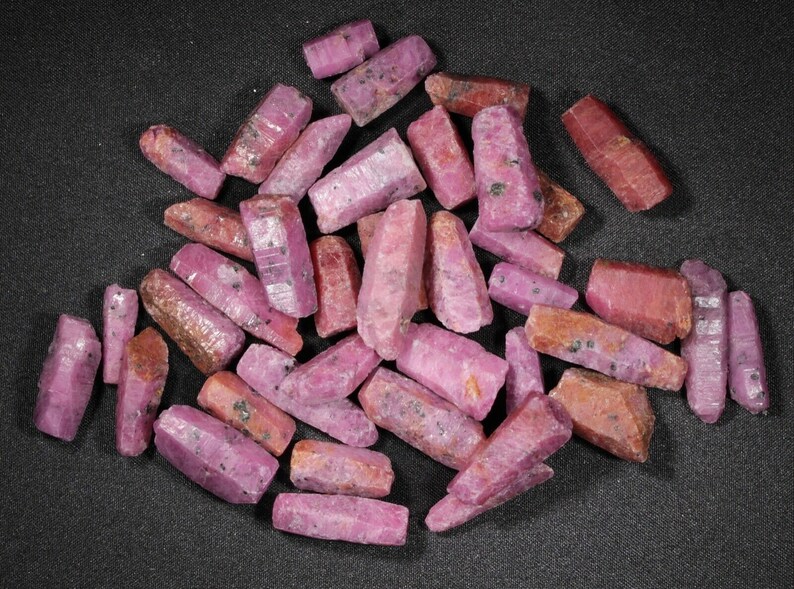 May include: A collection of pink ruby crystals, each with a pointed tip and a rough, natural texture. The crystals are arranged in a pile on a black background.