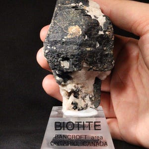 May include: A black biotite mineral specimen on a clear display stand. The label reads "BIOTITE, BANCROFT area, ONTARIO, CANADA".
