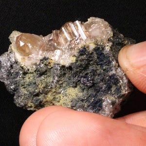 Puede incluir: Primer plano de un espécimen mineral con un grupo de cristales translúcidos de color marrón claro sobre una matriz oscura y moteada. Los cristales tienen una estructura prismática, y la matriz tiene una mezcla de tonos verde oscuro, grises y negros.
