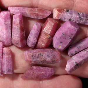 May include: A collection of pink ruby crystals, some with a darker purple hue, in various sizes and shapes. The crystals are rough and have a natural, unpolished look.