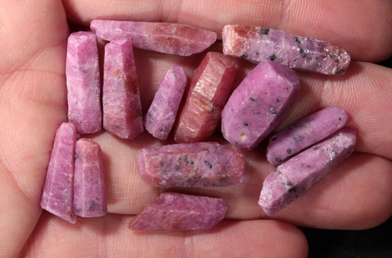 May include: A collection of pink ruby crystals, some with a darker purple hue, in various sizes and shapes. The crystals are rough and have a natural, unpolished look.