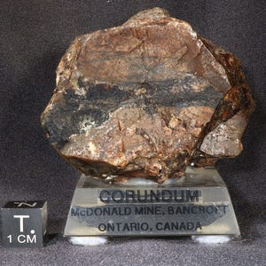 May include: A dark brown and black corundum rock specimen on a clear display stand. The label reads "CORUNDUM MCDONALD MINE, BANCROFT ONTARIO, CANADA".
