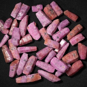 May include: A collection of pink ruby crystals, each with a unique shape and size. The crystals are arranged in a random pattern on a black background.
