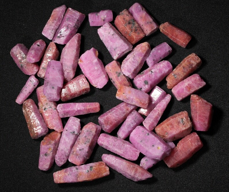 May include: A collection of pink ruby crystals, each with a unique shape and size. The crystals are arranged in a random pattern on a black background.