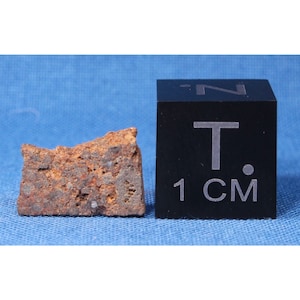Pode incluir: Um fragmento de meteorito marrom enferrujado com uma superfície áspera e irregular. O fragmento tem aproximadamente 1 centímetro de tamanho, como indicado pelo cubo de escala ao lado dele.