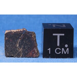 May include: A dark gray, irregularly shaped rock with a crack running through it. The rock is about 1 inch wide and is next to a black cube with the text "1 CM" on it.