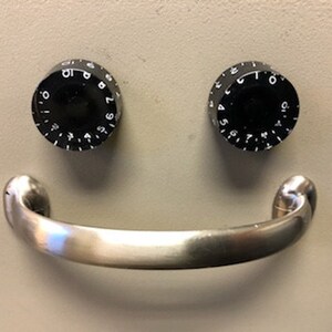 May include: A silver metal handle with two black round dials with numbers on them. The dials are positioned above the handle, creating a smiley face.