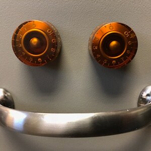 May include: A silver metal handle with two orange knobs with numbers on them. The knobs are positioned above the handle, creating a smiley face.