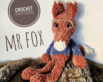 May include: A handmade crochet fox toy, wearing glasses and a blue and orange sweater. The fox has a white muzzle and ear tips. The text "CROCHET PATTERN" and "MR FOX" are in the background. The fox is sitting on a brown and white furry surface.