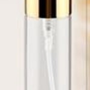 May include: A clear glass spray bottle with a gold-colored cap. The bottle has a long, thin, white plastic tube inside. The cap is a shiny, metallic gold color. The bottle is cylindrical and appears empty.