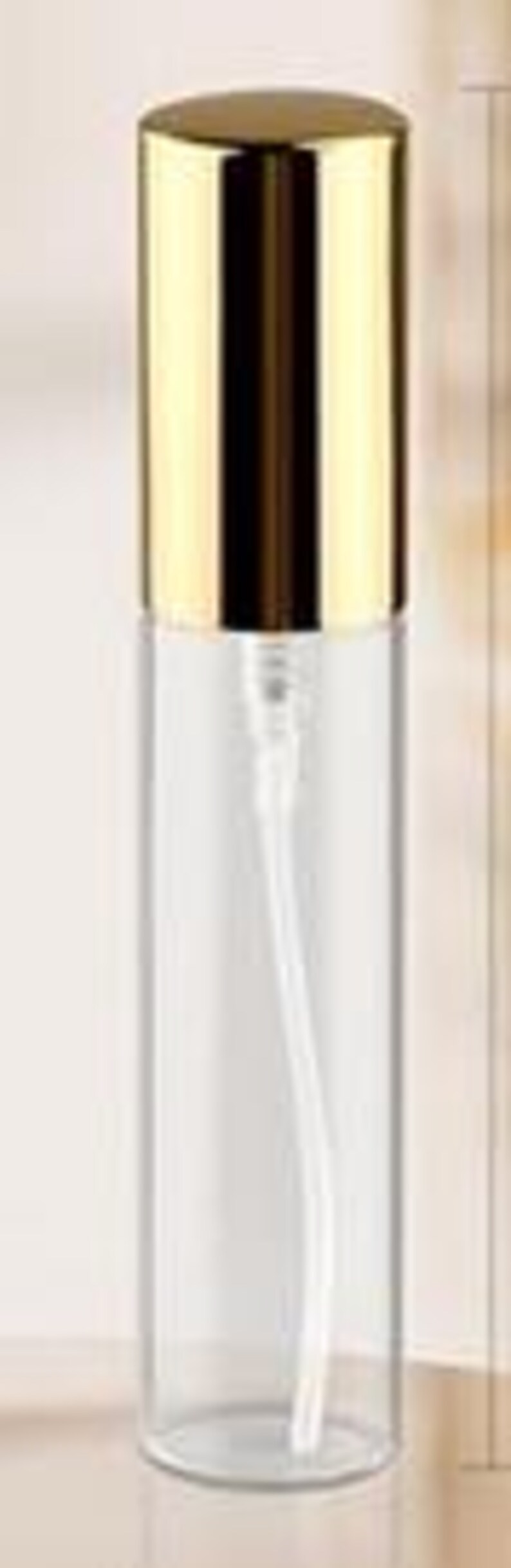 May include: A clear glass spray bottle with a gold-colored cap. The bottle has a long, thin, white plastic tube inside. The cap is a shiny, metallic gold color. The bottle is cylindrical and appears empty.
