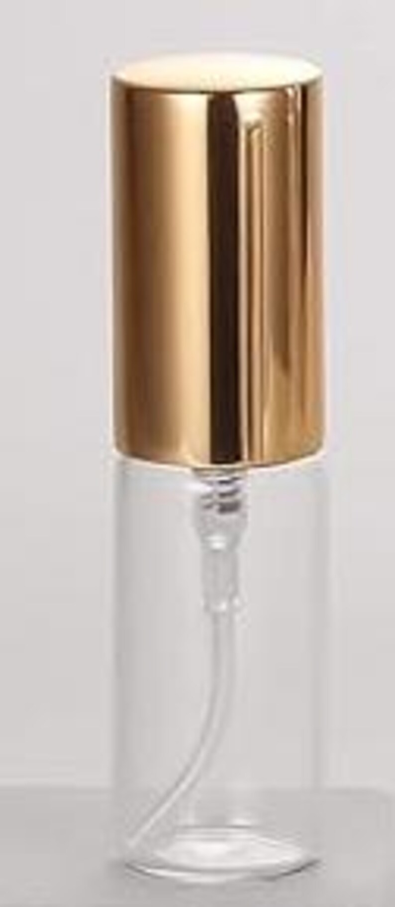 May include: A clear glass bottle with a gold-colored cap and a clear plastic pump. The bottle is cylindrical and appears to be empty. The cap is a shiny, metallic gold color. The pump extends into the bottle.