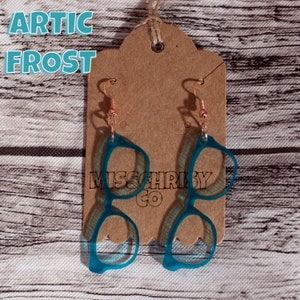 May include: Teal blue resin glasses shaped like eyeglasses dangle from rose gold hooks on a brown card with the text "Artic Frost" and "Miss Chrissy Co."