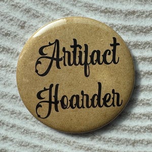 Op de afbeelding: Een ronde, bruinachtige knoop met de woorden "Artifact Hoarder" in zwart schrift. De knoop is gecentreerd op een getextureerd, lichtgekleurd oppervlak met gebogen lijnen.