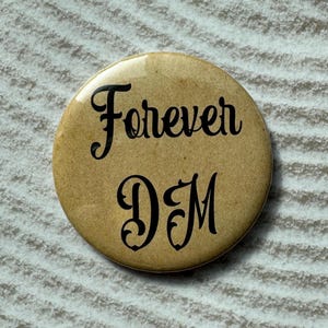 May include: A round, tan button with the words "Forever DM" in black script. The button is set against a textured, light gray background.