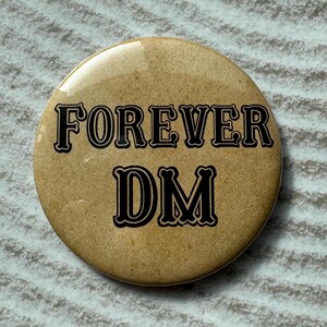 May include: A round, tan button with the words "FOREVER DM" in black, stylized lettering. The button has a glossy finish and is set against a textured, light gray background.