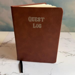 Op de afbeelding: Een bruin notitieboek met een gladde, matte kaft waarop de tekst "QUEST LOG" in een schreeflettertype op de voorzijde is gedrukt. Het notitieboek staat op een kier, waardoor crèmekleurige pagina's en een dun zwart leeslint zichtbaar zijn.