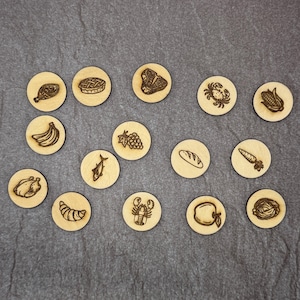 May include: A collection of fourteen circular light-colored wooden tokens, each featuring a dark laser-engraved illustration of a different food item, including a chicken leg, pie, steak, crab, corn, bananas, grapes, fish, bread, carrot, croissant, lobster, apple, and cabbage.