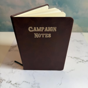 May include: A dark brown notebook with a smooth cover featuring the gold embossed text CAMPAIGN NOTES on the front. The notebook is slightly open, revealing several cream-colored pages inside, and includes a thin black ribbon bookmark.