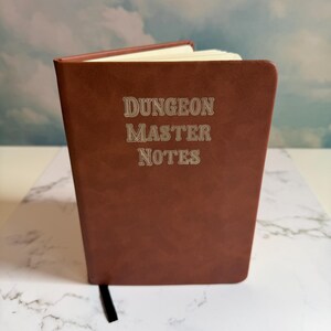 May include: A brown notebook with a smooth, matte finish featuring the text "DUNGEON MASTER NOTES" embossed in a serif font on the front cover. The notebook is slightly open, revealing white pages inside and a black ribbon bookmark at the bottom.