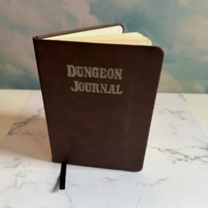 Op de afbeelding: Een donkerbruin dagboek met een gladde kaft waarop de tekst "DUNGEON JOURNAL" in een lichte kleur op de voorzijde is gedrukt. Het boek staat op een kier, waardoor crèmekleurige pagina's zichtbaar zijn, en bevat een dun zwart leeslint.