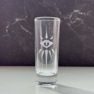 May include: Clear shot glass with a detailed etched design of an eye with radiating lines. The glass is cylindrical and stands upright, showcasing the intricate artwork. The background is a dark, marbled surface.