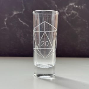 May include: A clear shot glass with a geometric design etched on the front. The design includes a large number 20 in the center. The glass is cylindrical with a flat base.