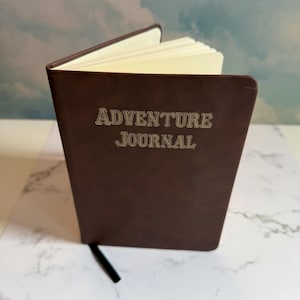 Op de afbeelding: Een donkerbruin dagboek met een gladde kaft voorzien van de in reliëf aangebrachte tekst ADVENTURE JOURNAL in een schreeflettertype. Het boek staat op een kier en toont crèmekleurige pagina's en een dun zwart lint als bladwijzer.