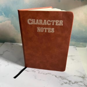 May include: A brown notebook with a smooth, leather-like cover featuring the text "CHARACTER NOTES" embossed in a light-colored, decorative font on the front. The notebook is standing upright, showing its rectangular shape and bound spine.