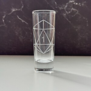 May include: A clear shot glass with a geometric design etched on the front. The design includes a large number 1 in the center. The glass is cylindrical and sits on a white surface with a dark background.