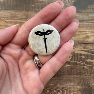 May include: A round button with a beige background and a black graphic of a sword with wings. The button is held in a hand.