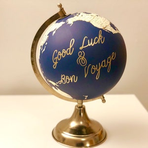 Może przedstawiać: Niebieski globus na złotej podstawie. Globus ma złoty napis "Good Luck & Bon Voyage".
