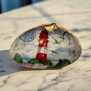 May include: A decorative seashell with a painted coastal scene. The artwork features a red and white lighthouse, seagulls, and a blue sky. The shell's edge is trimmed with gold.