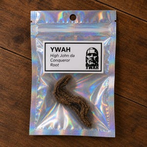 May include: A brown, textured High John da Conqueror root inside a holographic, iridescent plastic pouch. The white label features the text YWAH, High John da Conqueror Root, and a black and white graphic illustration of a human face.
