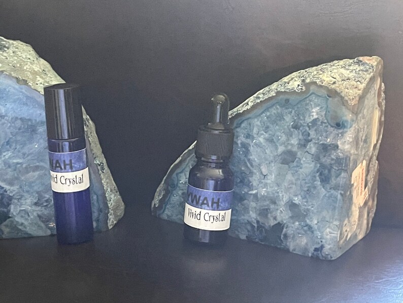 Puede incluir: Dos botellas de vidrio azul oscuro con tapas negras, etiquetadas como "YWAH Vivid Crystal", se exhiben frente a dos grandes rodajas de geoda azul-gris. Las botellas tienen un aplicador de bola y un cuentagotas. El fondo es oscuro.