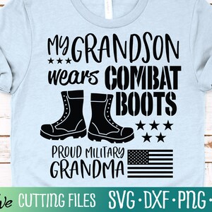 Pode incluir: Uma camiseta azul clara com texto preto que diz "My Grandson wears Combat Boots" e "Proud Military Grandma". O design também inclui um par de botas de combate pretas e uma pequena bandeira americana.