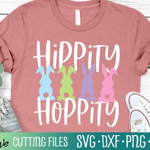 Może przedstawiać: Różowa koszulka z napisem "Hippity Hoppity" w kolorze białym. Jest na niej pięć pastelowych sylwet królików z sercami pod spodem.