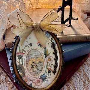 May include: An oval-shaped decorative frame featuring a gold-toned ribbon bow at the top. The center displays a botanical illustration of daisies and wildflowers surrounding a central oval window containing a pressed flower over handwritten script.
